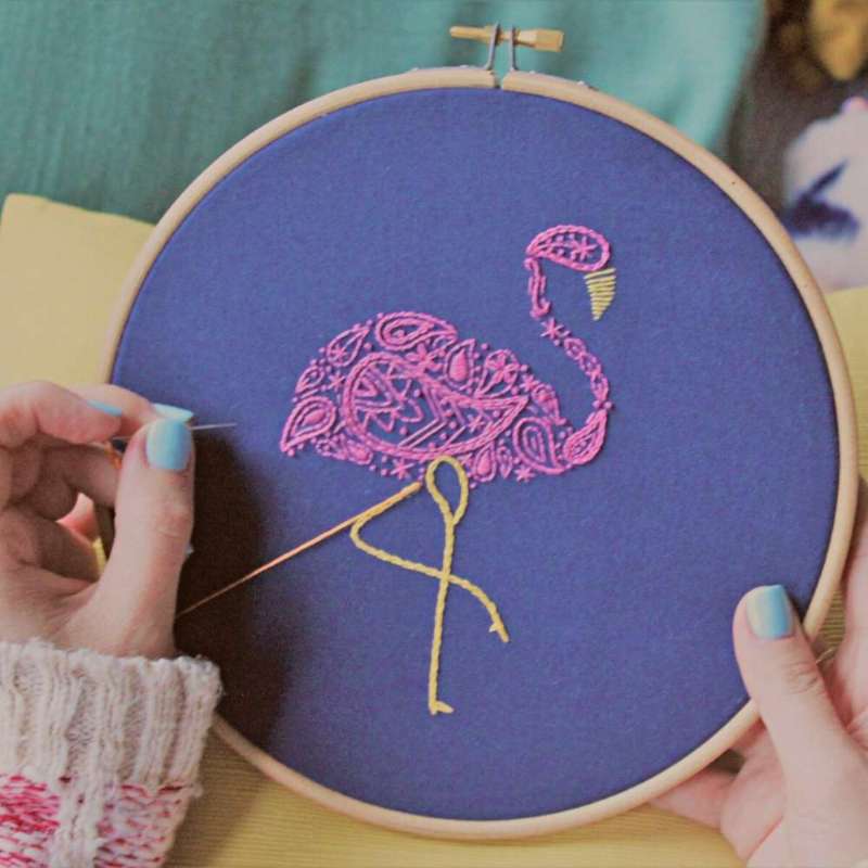 Embroidered pink flamingo being stitched on navy fabric with needle held in hand