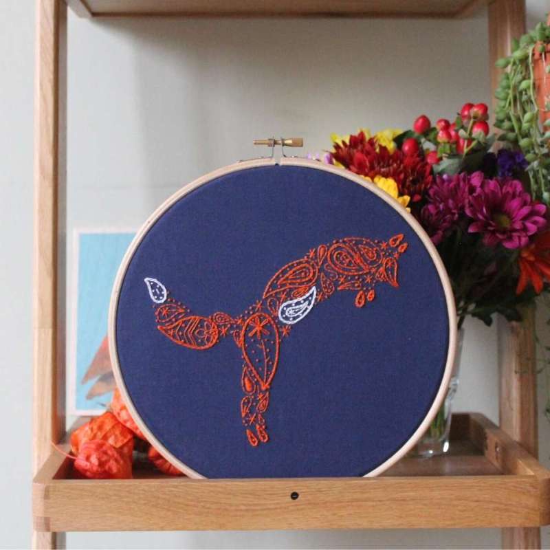 Orange and white pouncing fox embroidered on navy fabric in a wooden hoop