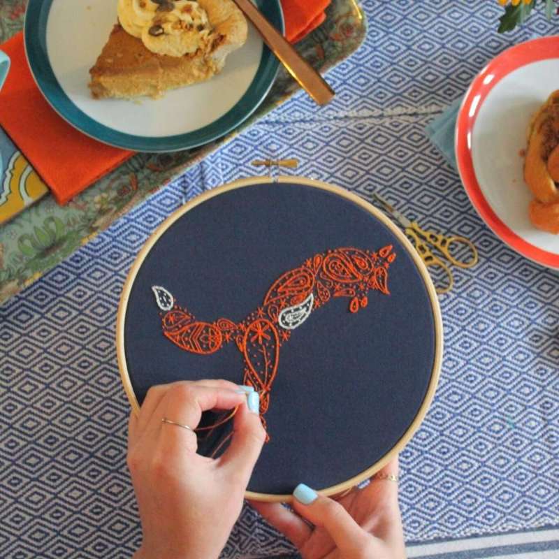 Orange and white fox embroidery kit being stitched on navy fabric with cakes on table in background