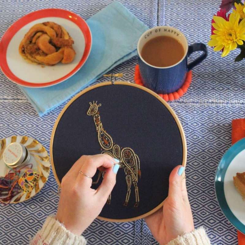 Hands embroidering a paisley giraffe embroidery design in shades of brown on navy fabric
