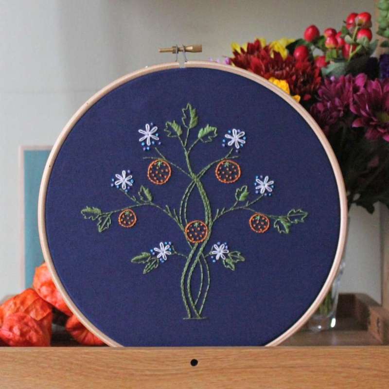May morris inspired orange tree embroidery design on navy fabric in wooden hoop on a shelf with flowers in background