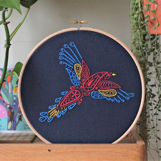 Red, yellow and blue embroidered parrot on navy fabric in wooden hoop rested on shelf