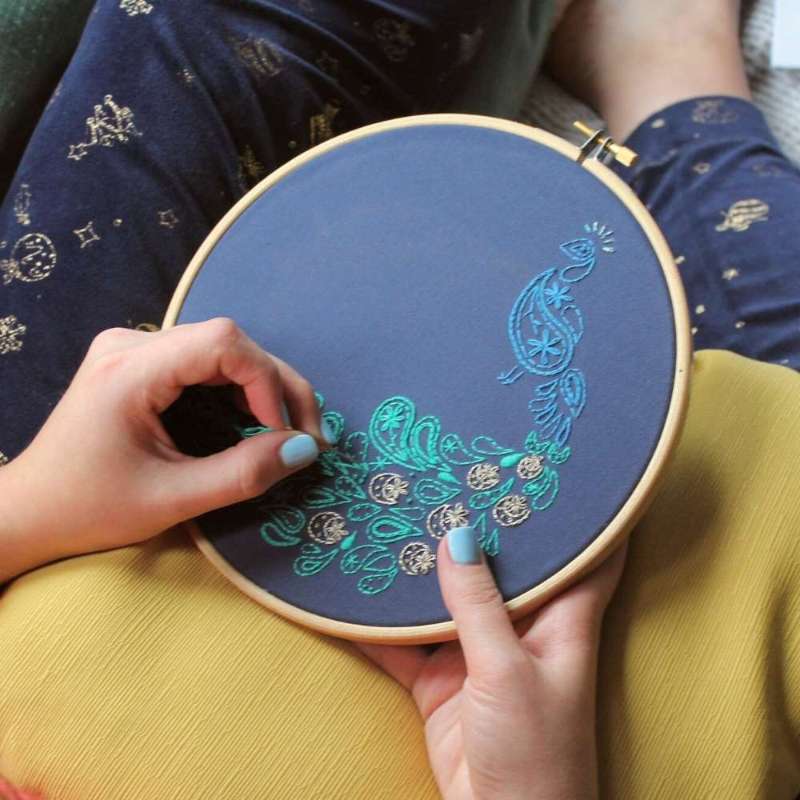 Blue, teal and gold paisley embroidered peacock on navy fabric held in hands on a sofa