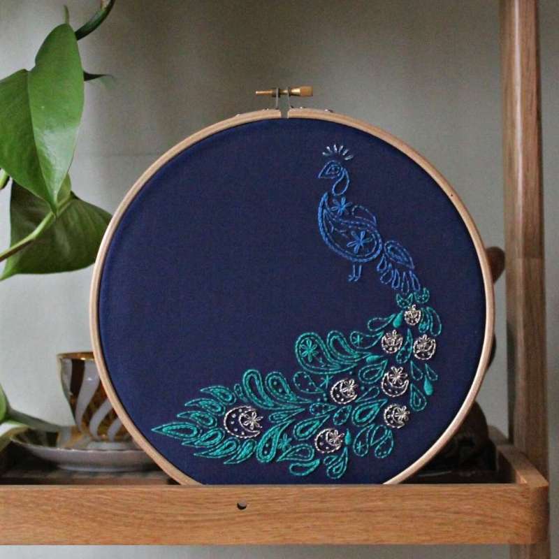 Paisley peacock embroidery kit shown completed on navy fabric in wooden hoop on a shelf