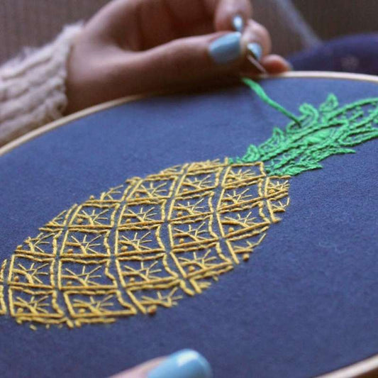 A yellow and green pineapple embroidered on navy fabric with green thread in a needle