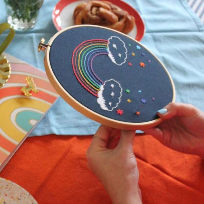 A side photo of someone embroidering a rainbow on navy blue fabric with cake in the background