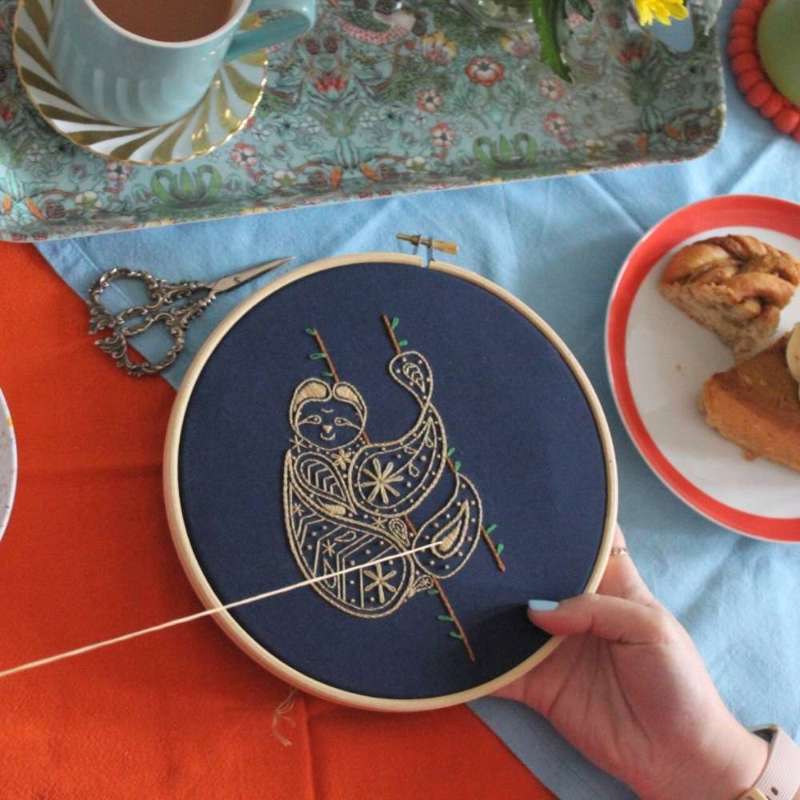 A hand holding a embroidery hoop with a sloth being embroidered on navy fabric