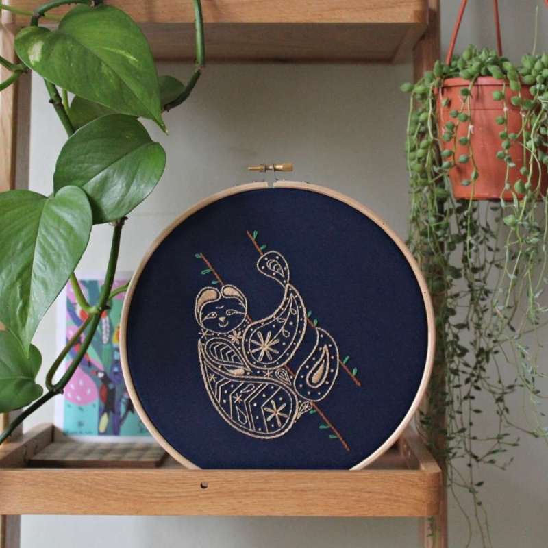 A brown sloth embroidered onto navy fabric in a wooden hoop on a shelf